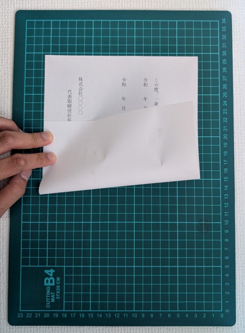 緑色のカッターマットの上で、退職届の下側を持ち上げて下から折りたたんでいる様子。縦書きの文面が上部に見えている。